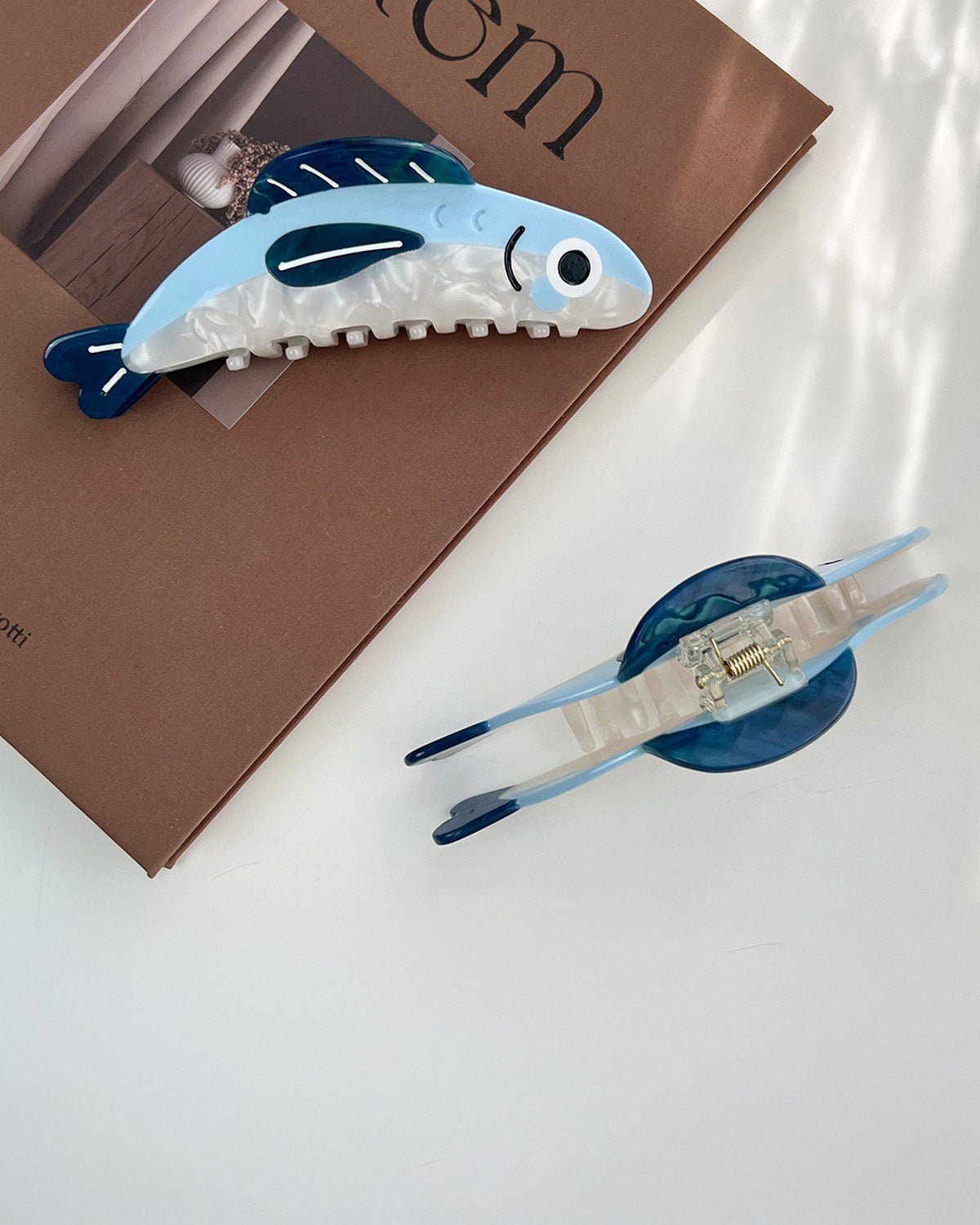 Two blue and white fish-shaped hair clips on a white surface with a brown book in the background