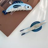 Two blue and white fish-shaped hair clips on a white surface with a brown book in the background