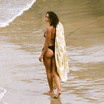 Woman in a black bikini with a towel draped over her shoulder walking on a beach
