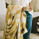Person holding a cup wrapped in a yellow and white patterned blanket on a couch
