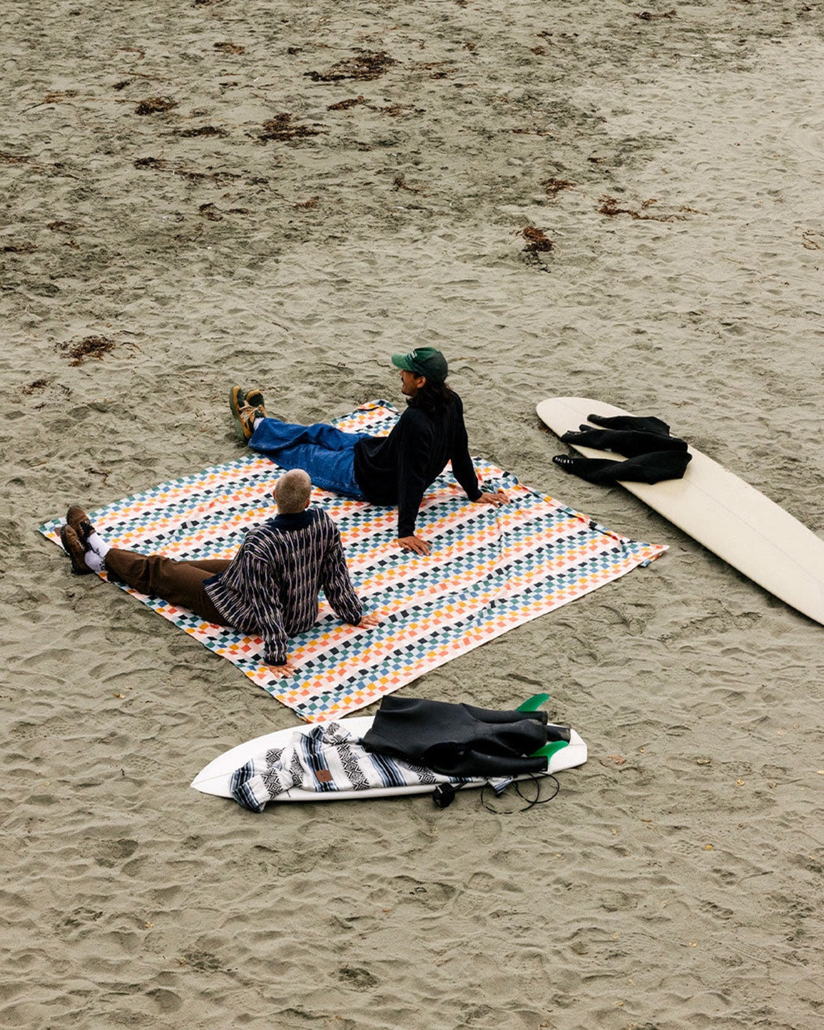 Two people sitting on a colorful blanket on a sandy beach with surfboards nearby