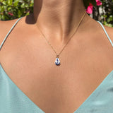 Necklace with a blue pendant worn by a person in a light blue top, with greenery in the background.