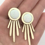 Gold earrings with opal stones held in a hand against a neutral background