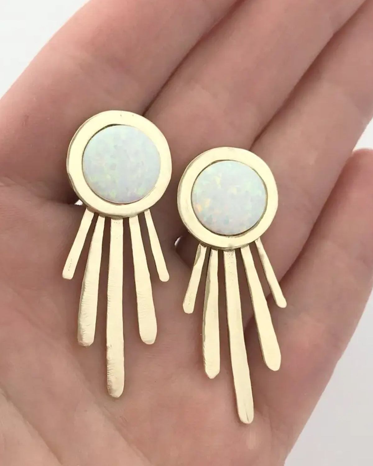 Gold earrings with opal stones held in a hand against a neutral background