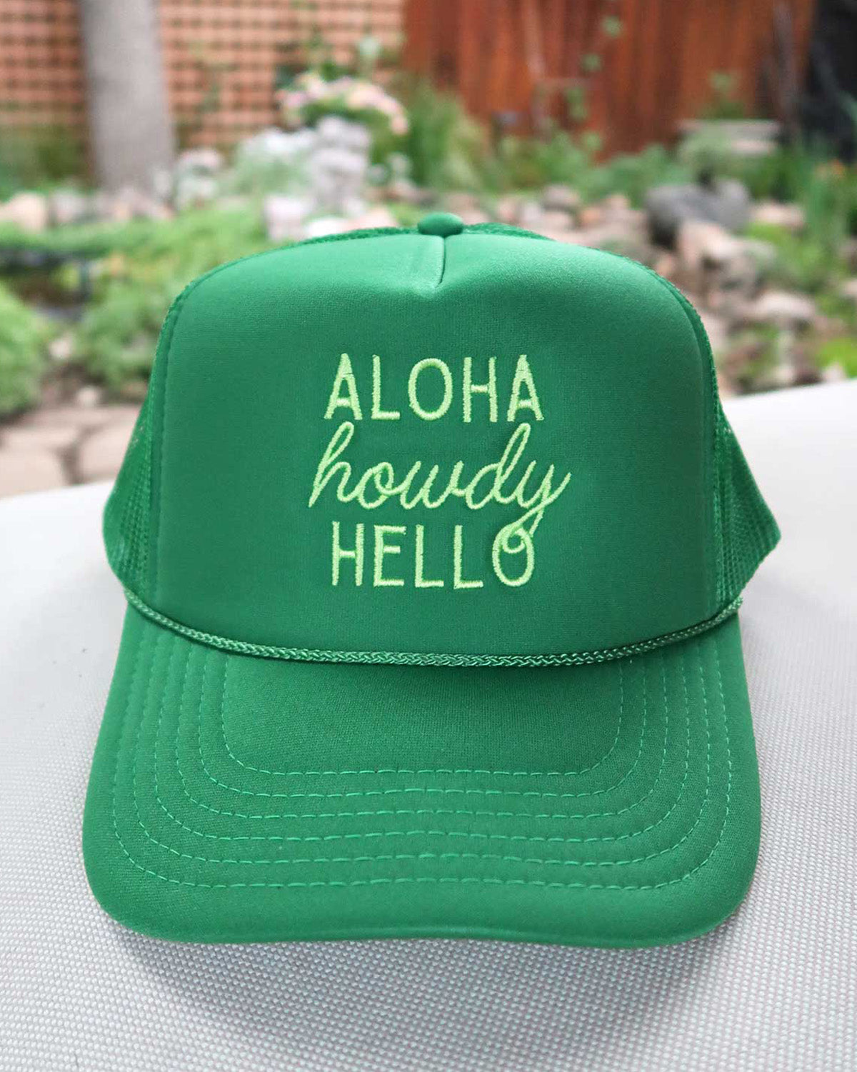 Green cap with 'Aloha howdy HELLO' text on a blurred natural background