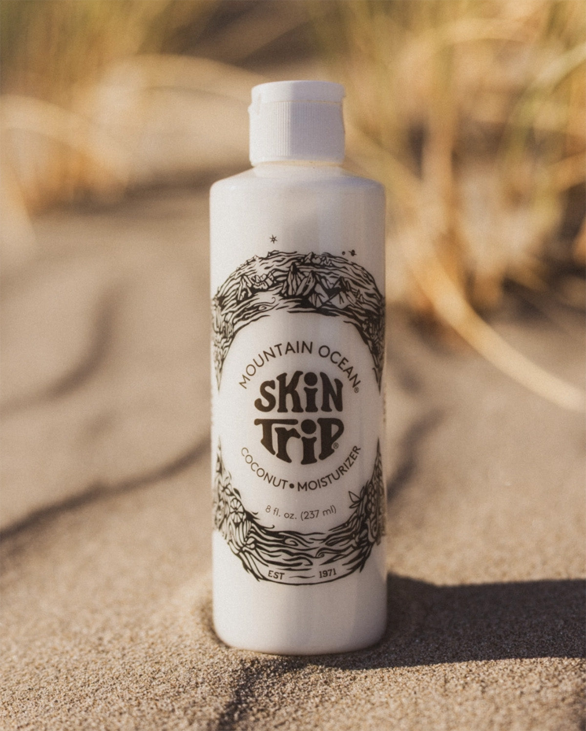 White bottle of Mountain Ocean Skin Trip product on sand with blurred natural background