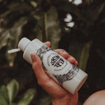 Hand holding a white bottle with black text and design against a blurred natural background